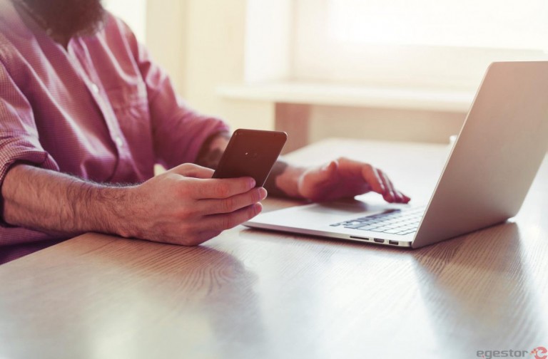 Homem de negócios conferindo a situação dos boletos emitidos em seu celular, enquanto navega no sistema de emissão no computador