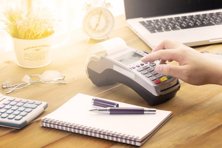 Close-up de um caderninho de anotações com uma caneta e dois tipos de calculadoras sendo usadas para descobrir os impostos da nota fiscal