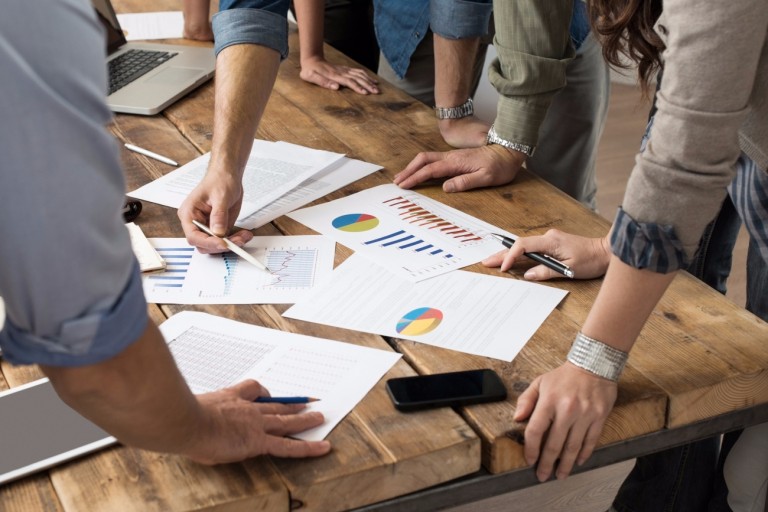 Ao redor de uma mesa de reuniões cheia de relatórios, várias pessoas conversam sobre os desafios que as microempresas enfrentam