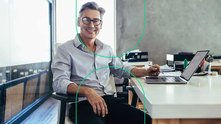 Gestor em seu escritório com o notebook aberto em sua mesa, olhando para a câmera e sorrindo