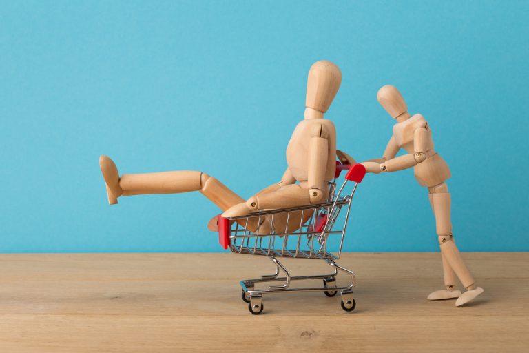 Dois bonequinhos de madeira: um sentado em um carrinho de mercado enquanto o outro empurra o carrinho