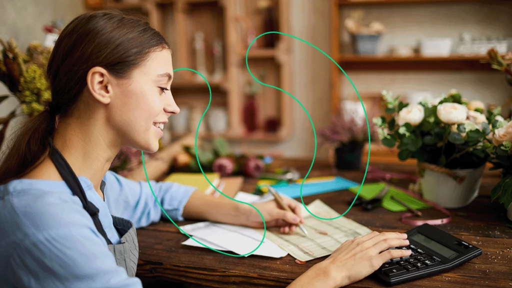 Microempreendedora no balcão de sua loja, usando uma planilha em papel e uma calculadora para fazer o financeiro do seu negócio