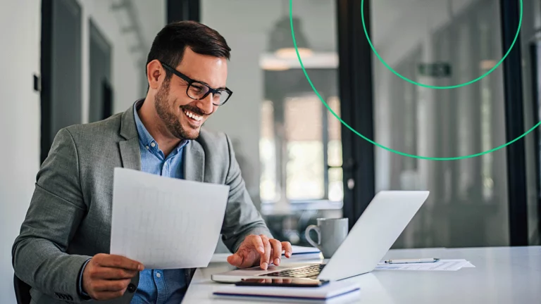 Empresário sorrindo enquanto segura um relatório na mão e digita no notebook, fazendo um Pix com segurança