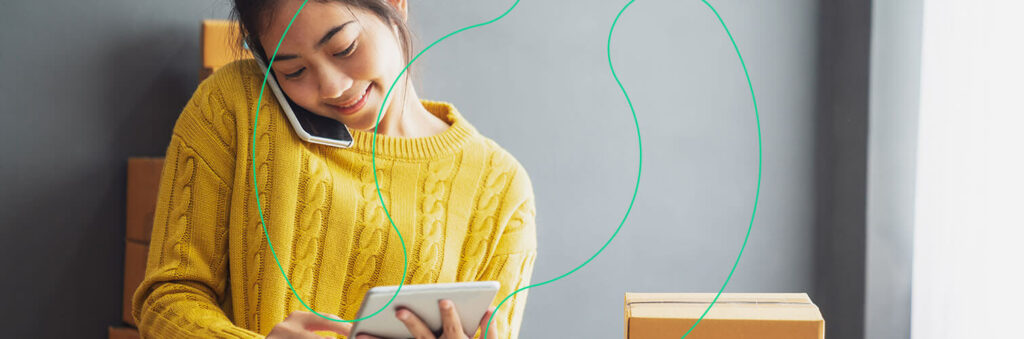Mulher sorrindo ao falar no telefone, ao mesmo tempo em que utiliza um tablet para conferir as entregas da Estoca